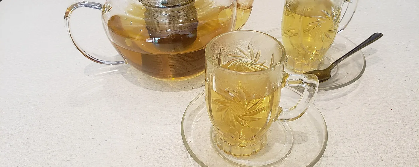 Glass teapot of pale peppermint tea with metal infuser, beside two etched glass cups on a white table