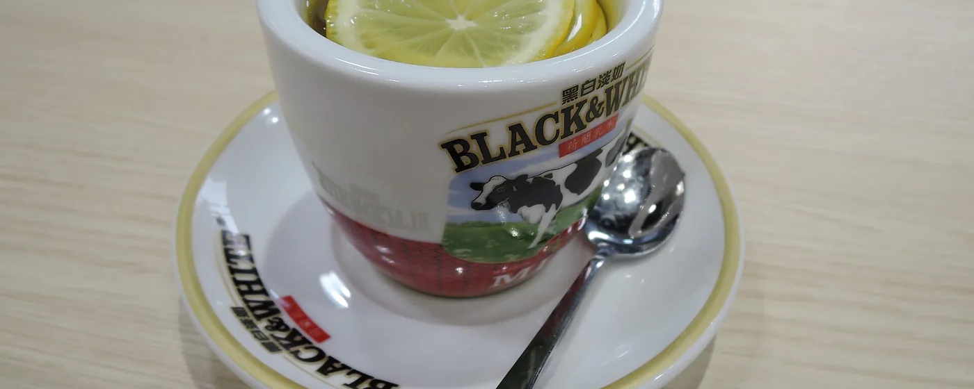 Hot lemon tea in a white cup and saucer with a lemon slice floating on top and a silver spoon alongside