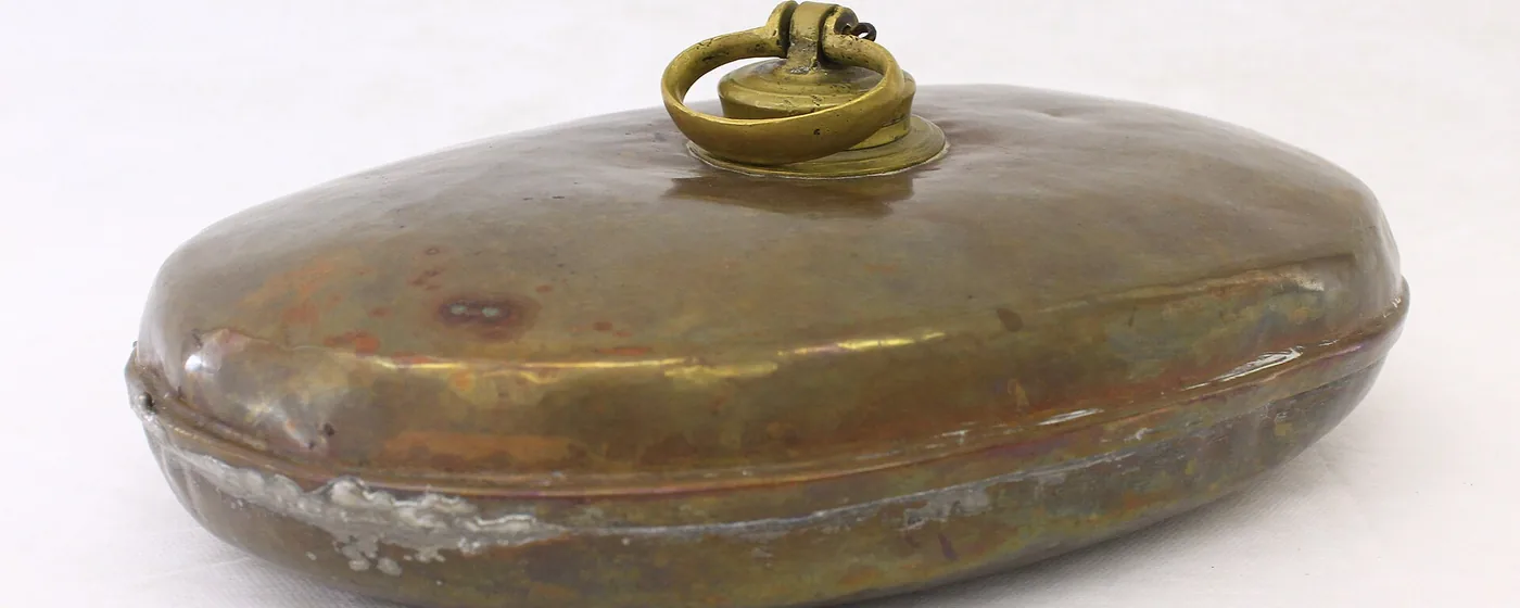 Vintage brass hot water bottle with a ring-topped screw cap, resting on a plain white background
