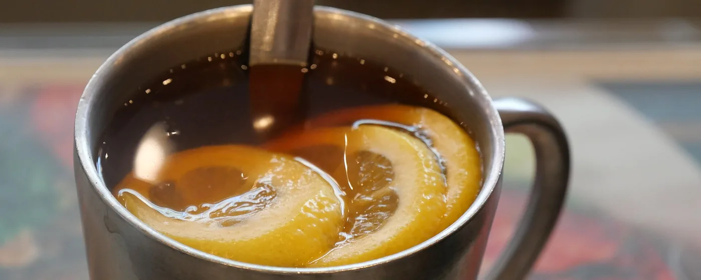 Close-up of a steel mug of amber lemon tea with two floating lemon slices and a spoon