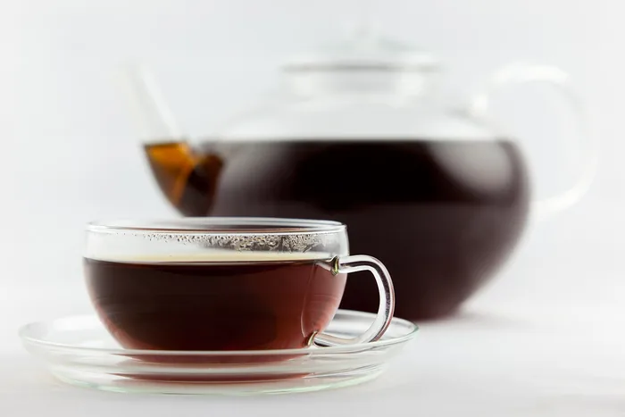 Glass teacup of amber Earl Grey tea on a saucer with a glass teapot of brewed tea behind, on white