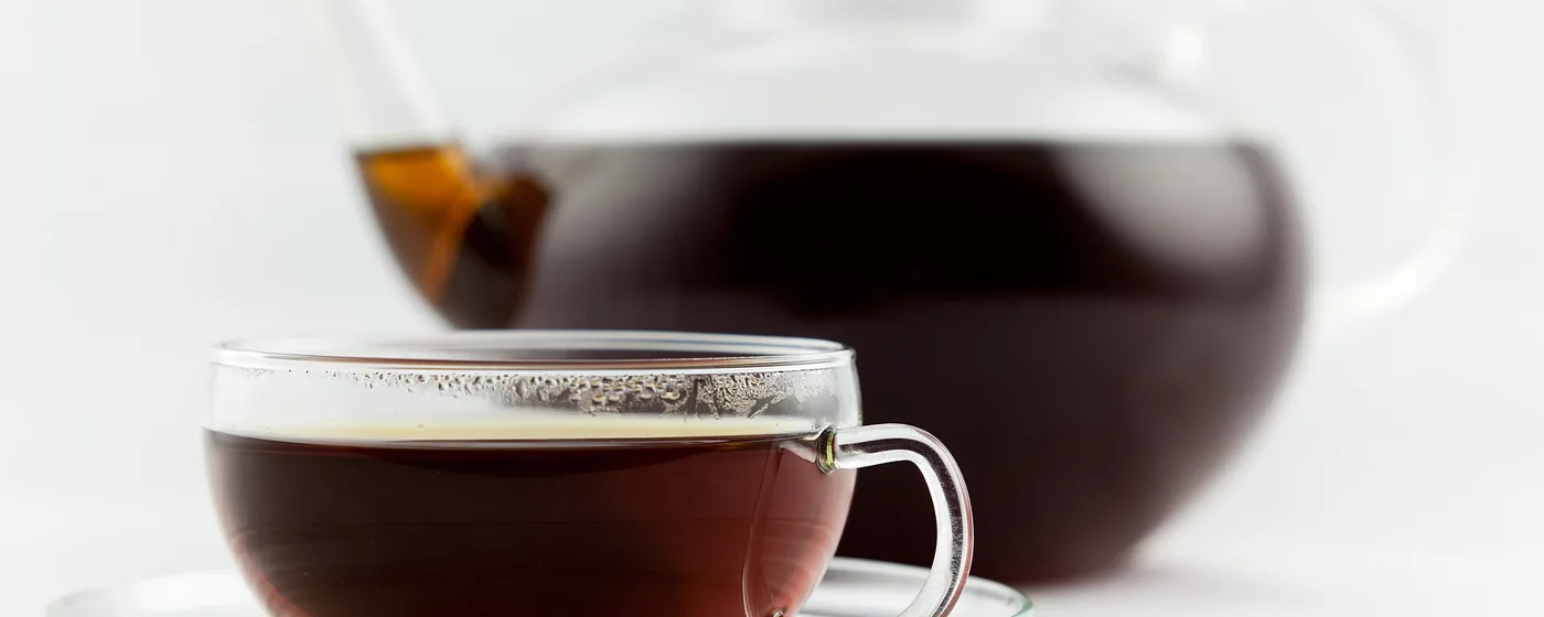 Glass teacup of amber Earl Grey tea on a saucer with a glass teapot of brewed tea behind, on white
