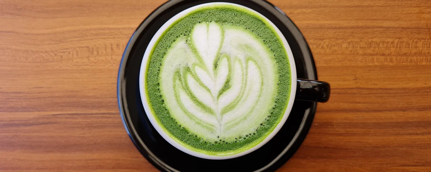 Overhead view of a bright green matcha latte with white rosetta latte art in a black cup on wood