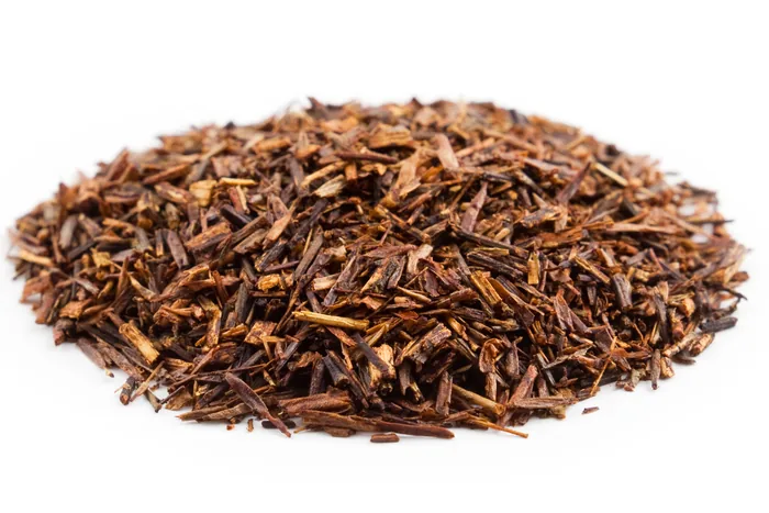 Heap of dried cut rooibos tea, reddish-brown needle-like leaf pieces, on a white background